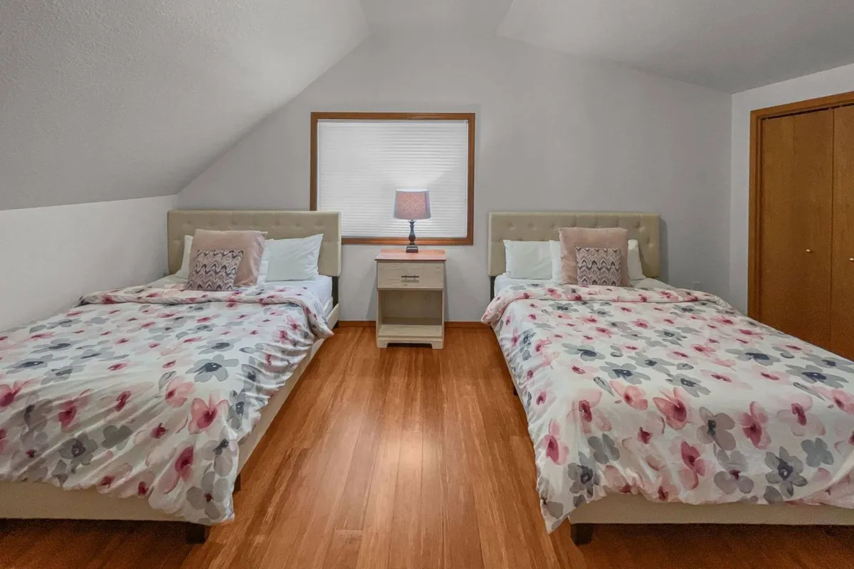 A spacious bedroom with two queen beds adorned in floral bedding, a nightstand with a lamp between them, and natural light streaming in through a window. The room feels airy and serene, ideal for families or groups.