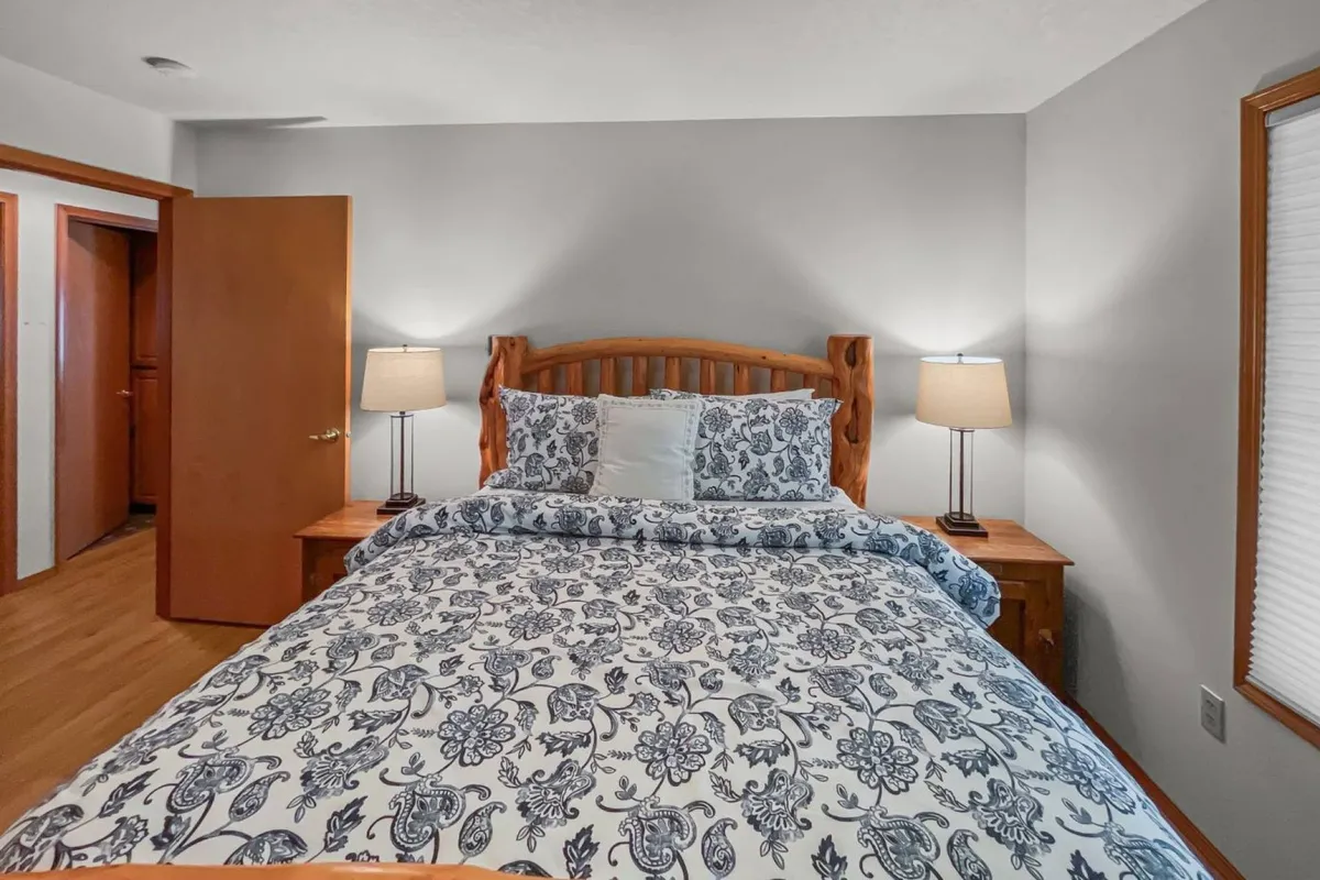 A bedroom featuring a queen bed with patterned bedding, a wooden headboard, and a nightstand with a lamp. Soft lighting and cozy textures create an inviting, restful atmosphere.