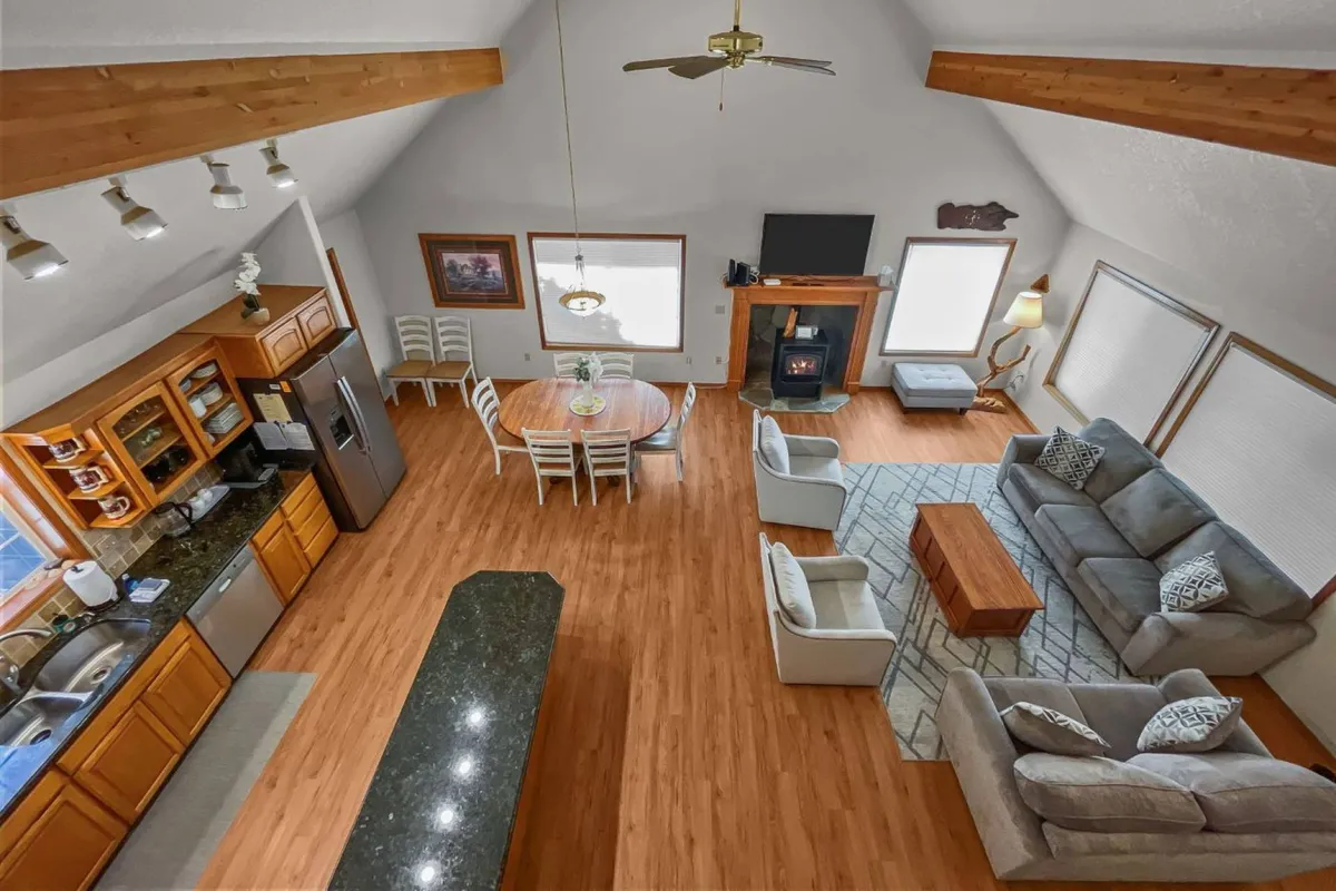 A spacious, open-concept living and dining area with a kitchen island, dining table, sofa seating area, and a cozy fireplace. The space is filled with natural light from large windows, highlighting vaulted ceilings and plush seating arrangements ideal for gatherings.