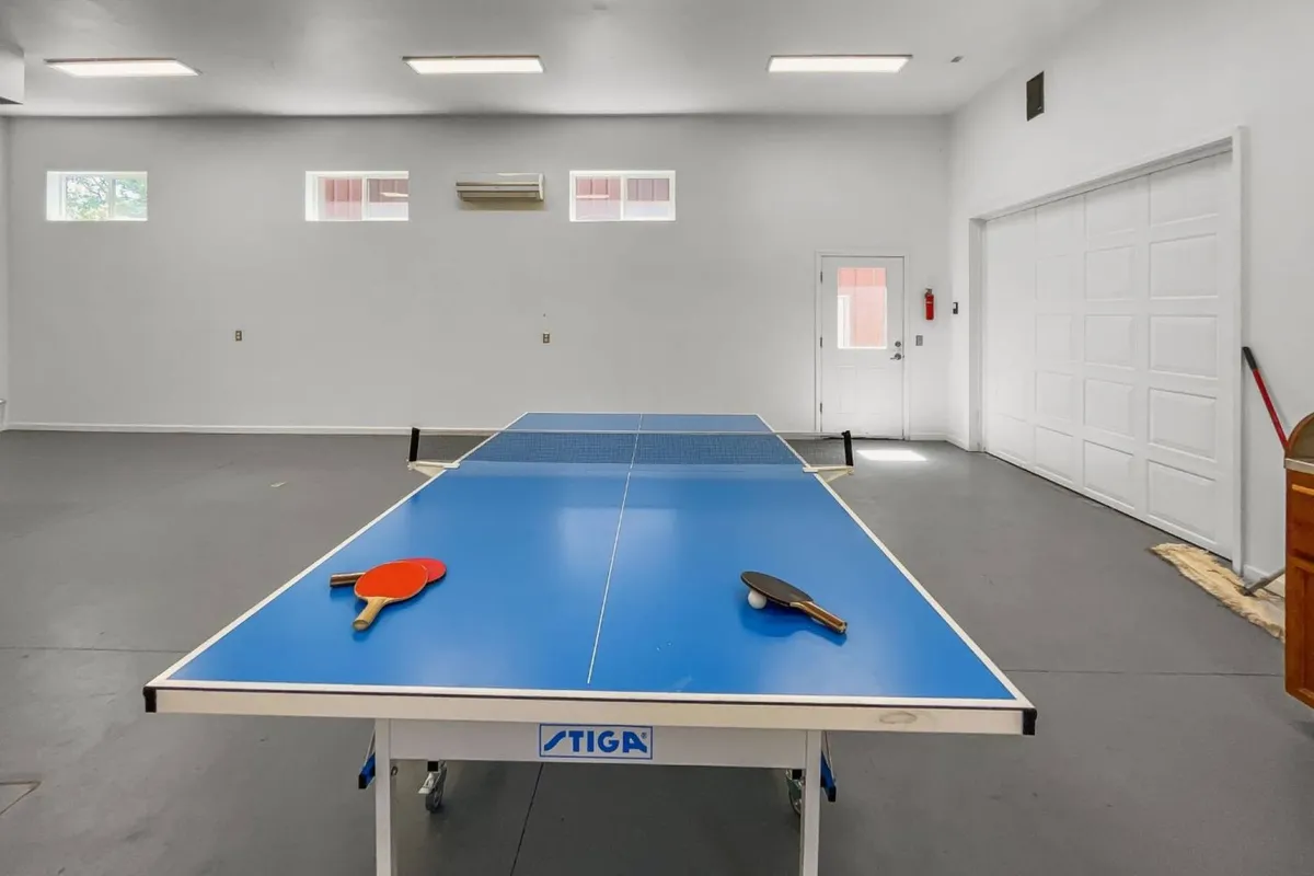 The image focuses on the ping pong table prominently placed within a converted garage space. Visible are paddles, the garage door, and windows that let in natural light, adding to the inviting and playful environment. The setup encourages both casual matches and spirited tournaments.