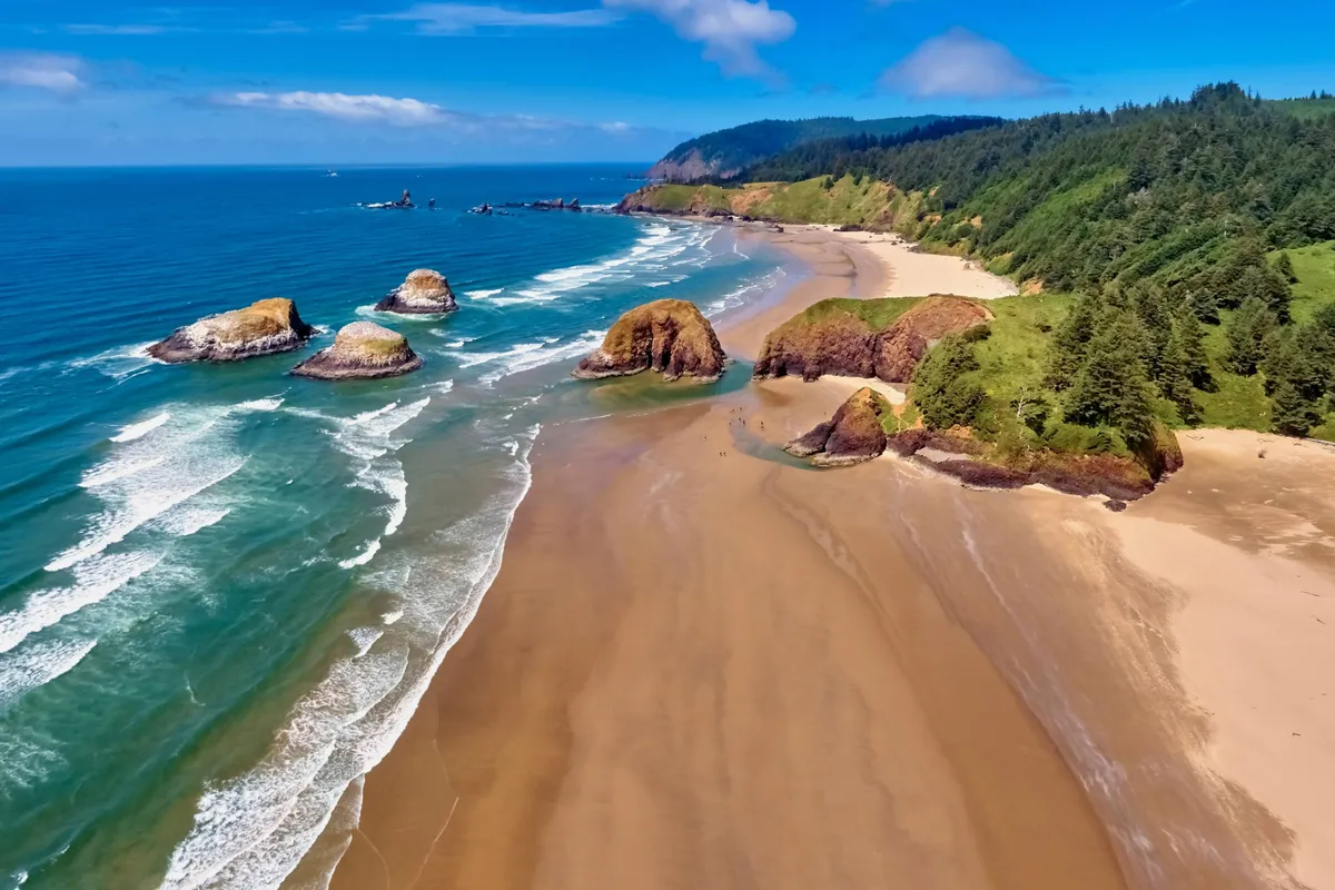 A scenic exterior view captures the natural beauty of the Oregon coast with dramatic rock formations, a sandy beach, ocean waves, and a lush forest backdrop. The image invites thoughts of beach walks, exploring tide pools, and picnicking, connecting the property to nearby attractions like Oceanside and Rockaway Beach.