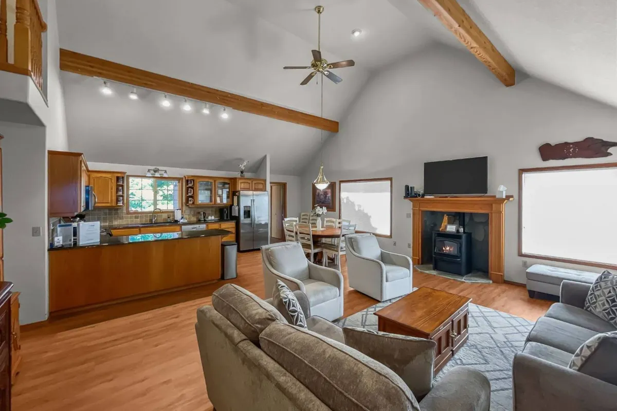 A welcoming view of the living space featuring a plush sofa, adjacent dining area, kitchen island, and a glowing fireplace. The room is bright with natural light, accentuating the comfortable seating and open flow between spaces.