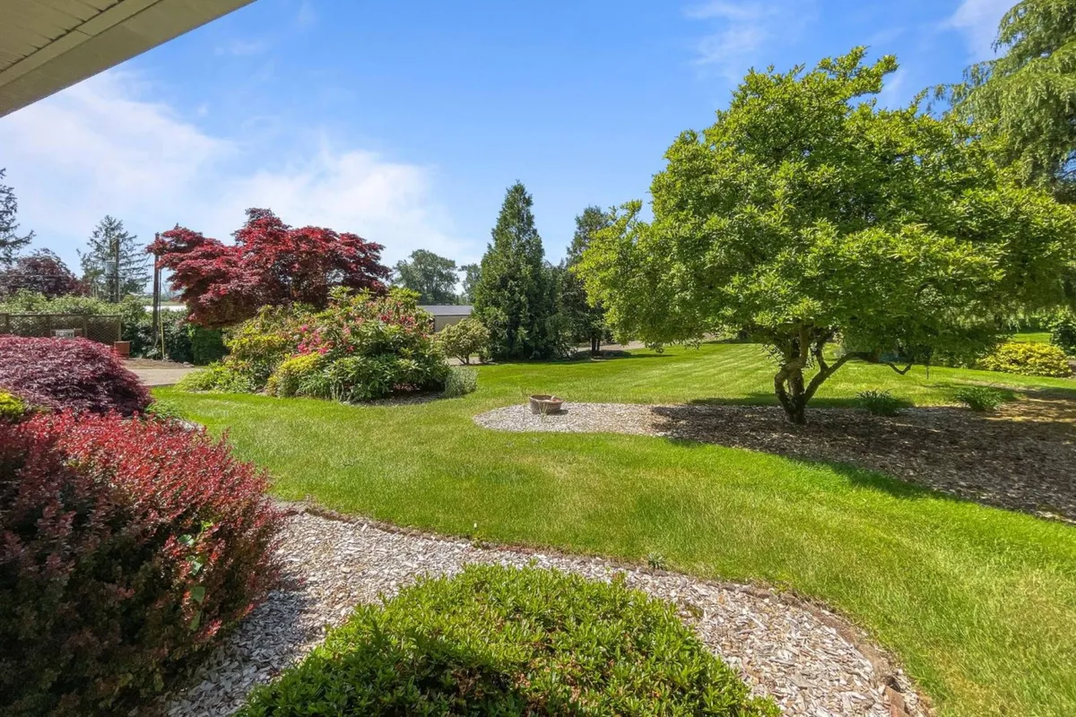 The image showcases a lush green lawn framed by vibrant flower beds and mature shade trees, with winding decorative stone pathways inviting exploration. The expansive, beautifully landscaped garden promises both sun-dappled spaces and cool, serene spots beneath the trees, all set against the peaceful backdrop of Oregon's natural beauty.