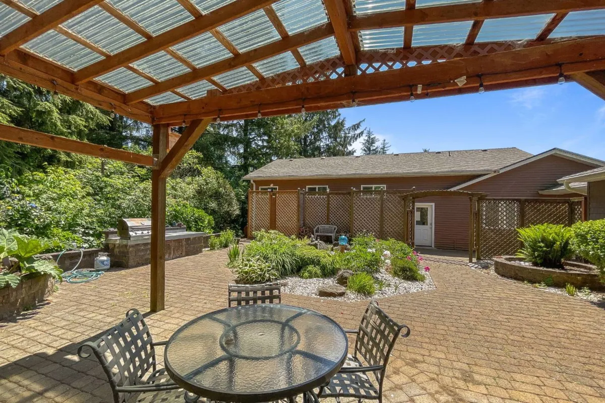 This image features an inviting outdoor dining table set beneath a pergola, surrounded by lush, landscaped gardens and a barbecue grill. The area exudes a relaxed, social vibe, ideal for gatherings and leisurely afternoons outdoors.